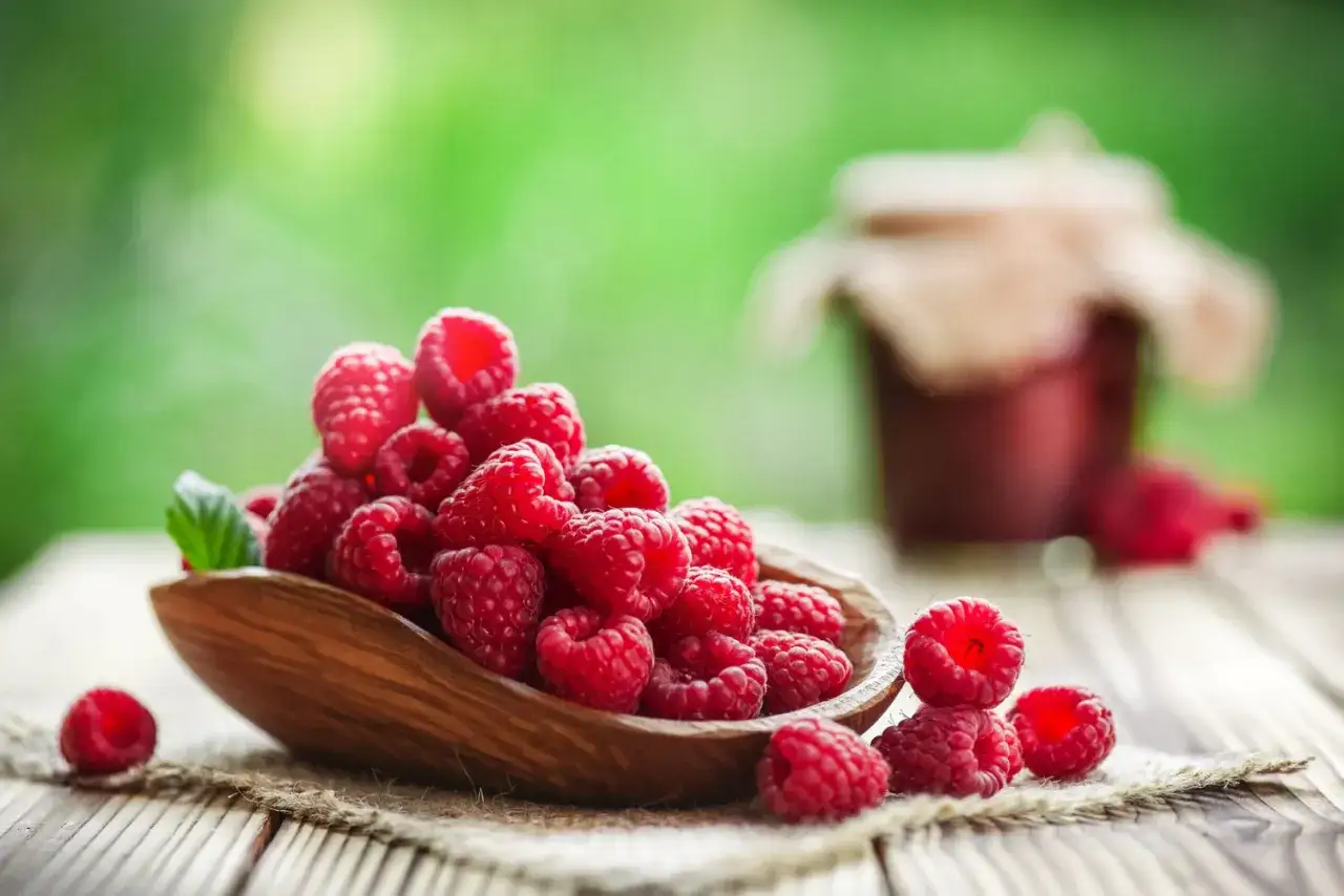 Frische Himbeeren in einer Holzschale, ein Symbol für gesunde Ernährung. Im Hintergrund ein Glas Marmelade.