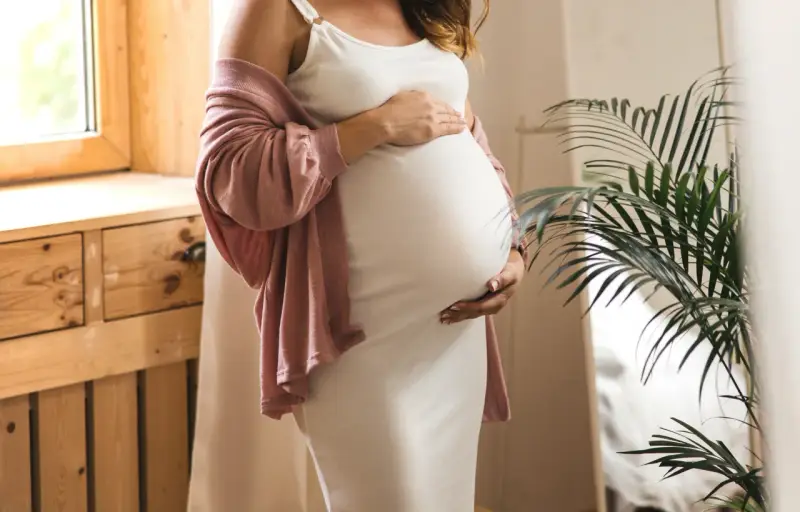 Frau in weißem Kleid und rosafarbenem Cardigan hält ihren Bauch, 34 ssw. Ein Fenster und eine Pflanze sind im Hintergrund.